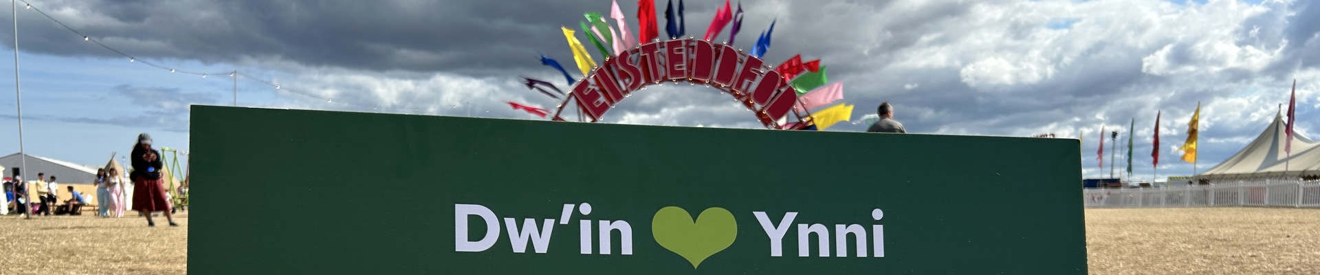 Arwydd yn darllen 'Dwi'n caru ynni adnewyddadwy i Gymru', gyda logo Trydan. Yn y cefndir mae arwydd Eisteddfod ar y maes.